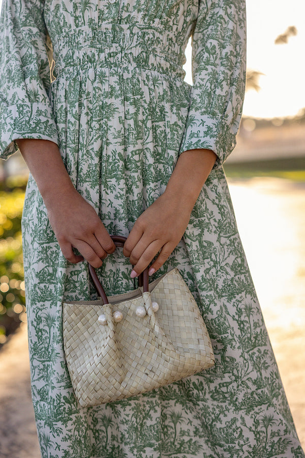 close up of a woman in a UPF 50+ green floral maxi dress with sun protection holding a straw handbag standing on a grassy lawn at a country club 
